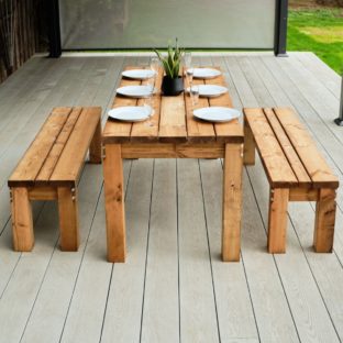 A chunky wooden rectangular outdoor dining table and 2 benches end of view on a grey deck with table set for dinner
