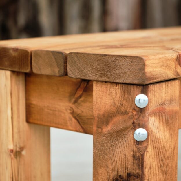 A close up of the end of a wooden backless outdoor bench showing chunky wooden planks joined with a robust metal bolt