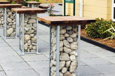 A gabion cage pedestal poseur height outdoor table with the cage filled with cotswold stone rocks and a square wooden table top located on a hotel patio