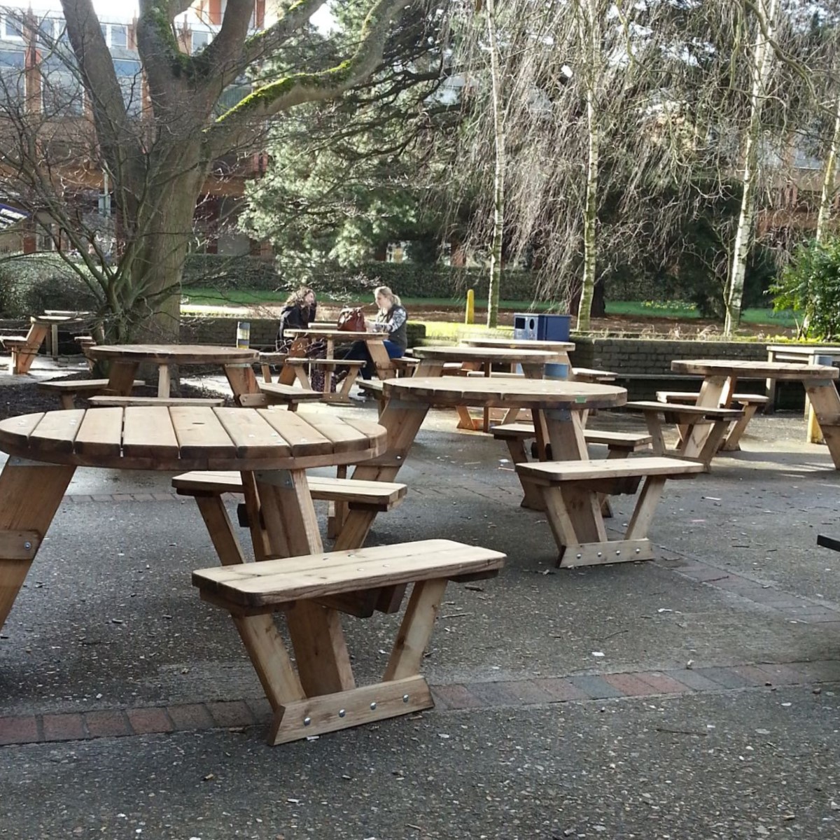 A patio area at Reading University with 8 circular wooden 6-seater picnic tables on and trees in the background