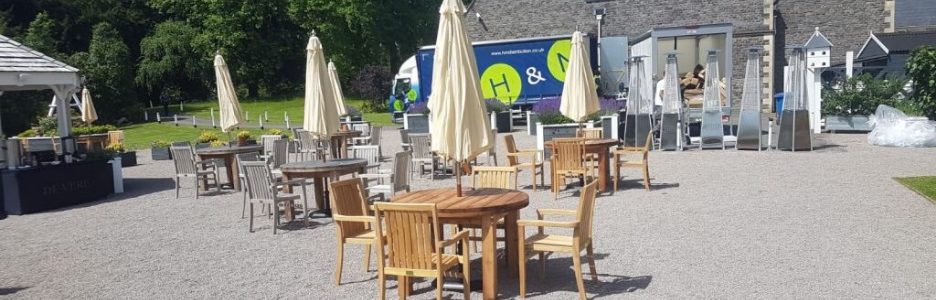 Teak outdoor dining tables chairs and parasols on a gravel terrace with a delivery van unloading in the background