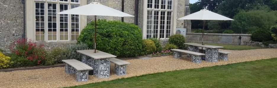 Outdoor rectangular dining tables with wooden tops and stone filled cages as legs situated on gravel at the front of the Horsley Estate Devere Hotel