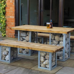 A rectangular garden table and 2 benches located on a patio. The table and benches have large cobble stone filled gabion legs and a wooden top