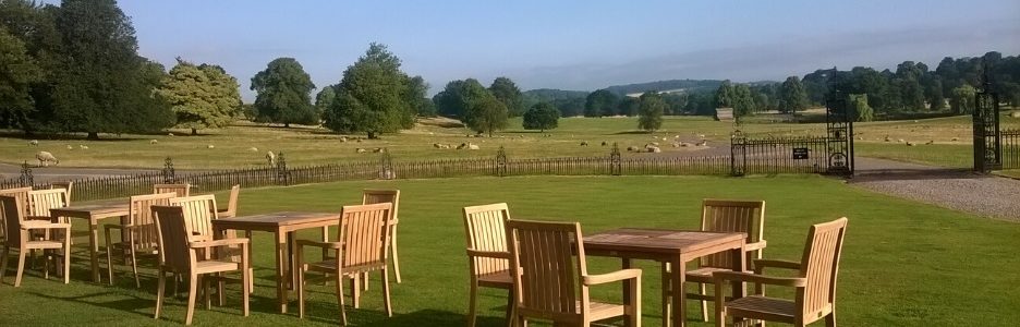 Three teak outdoor dining tables with four chairs around each one on a lawn overlooking a field of sheep