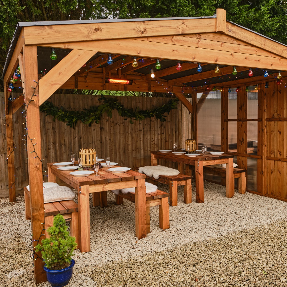 A square wooden gazebo with apex waterproof roof with 2 rectangular wooden tables underneath seating 12 dressed for christmas dinner