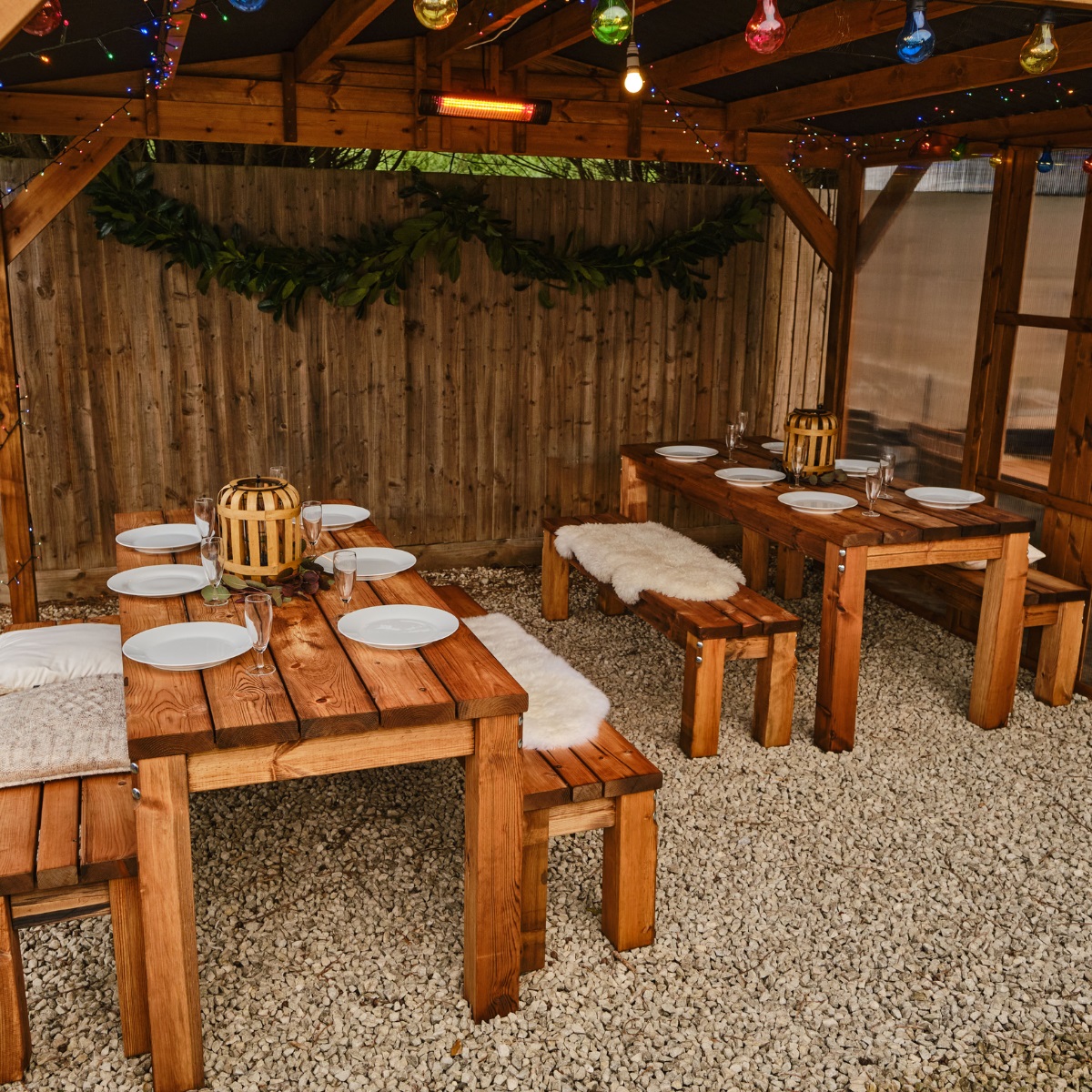 Two wooden rectangular outdoor tables with 2 bench seats either side with tables set for dinner underneath a wooden gazebo on a gravel patio
