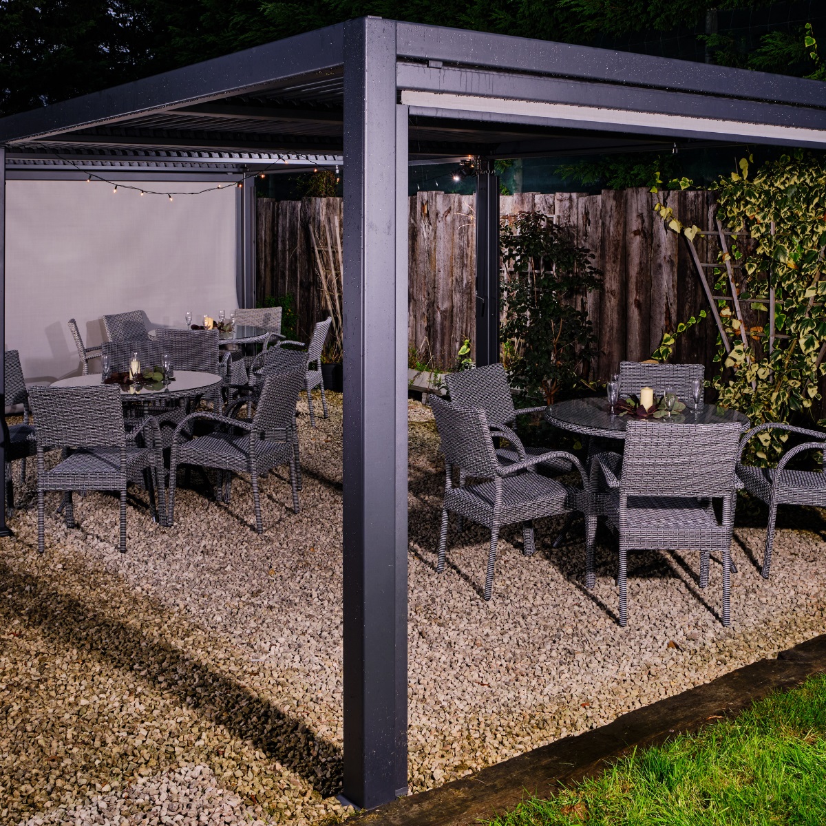 A dark grey metal rectangular weather proof gazebo with a flat louvered panel roof and 3 grey rattan dining tables and chairs underneath