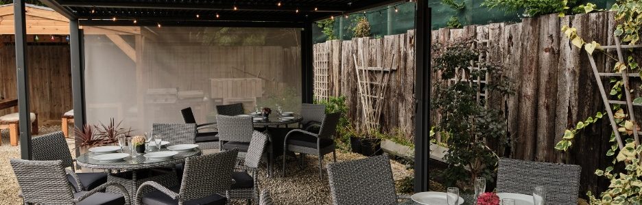 A close up of 3 round grey rattan tables and chairs sets underneath a grey metal gazebo with a flat roof on a gravel patio