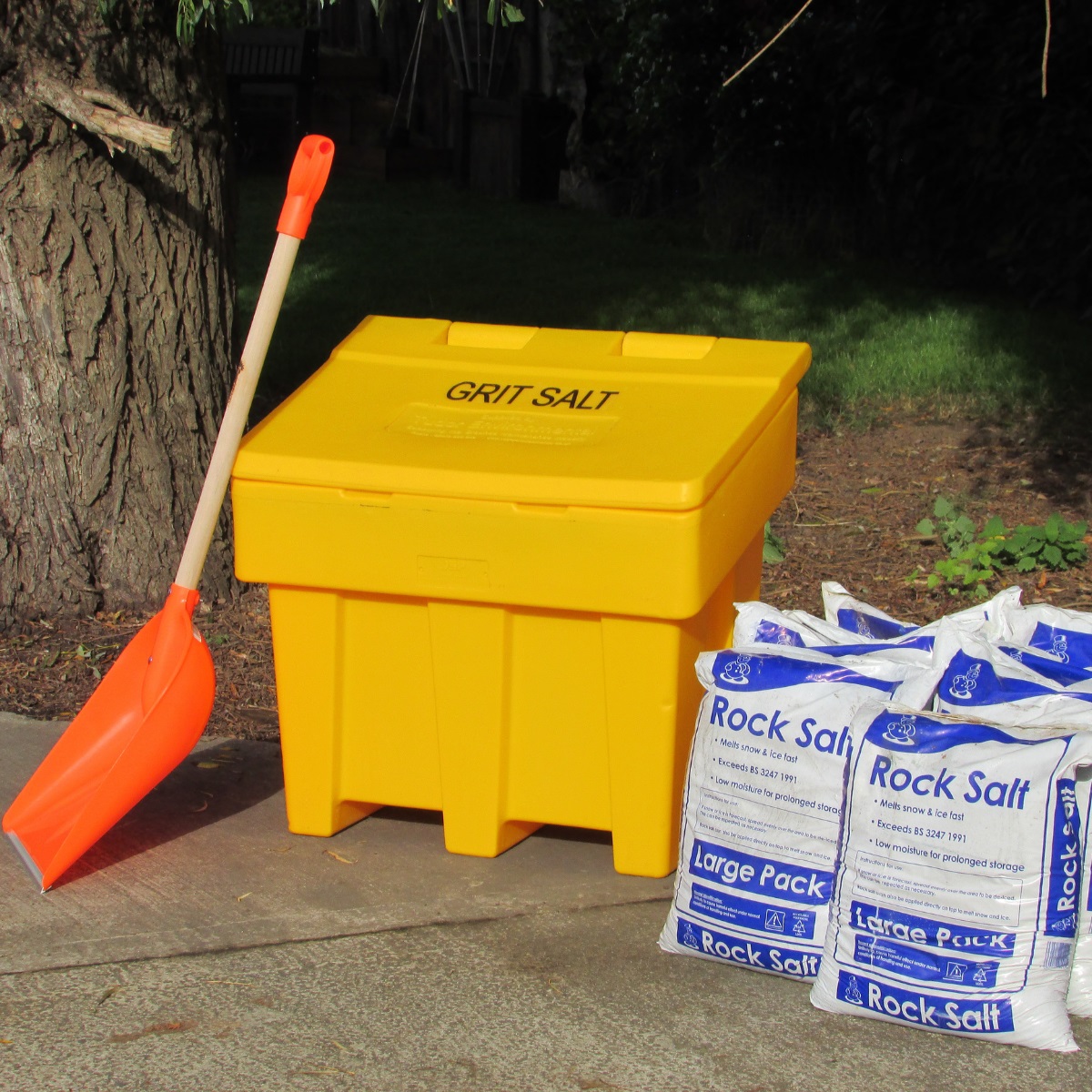 An orange snow shovel propped against a yellow grit bin with 6 bags of rock salt next to it
