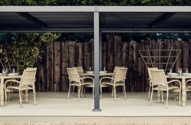 A dark grey metal rectangular gazebo with a flat slatted waterproof roof on a light grey deck with grey rattan tables and chairs arranged on it