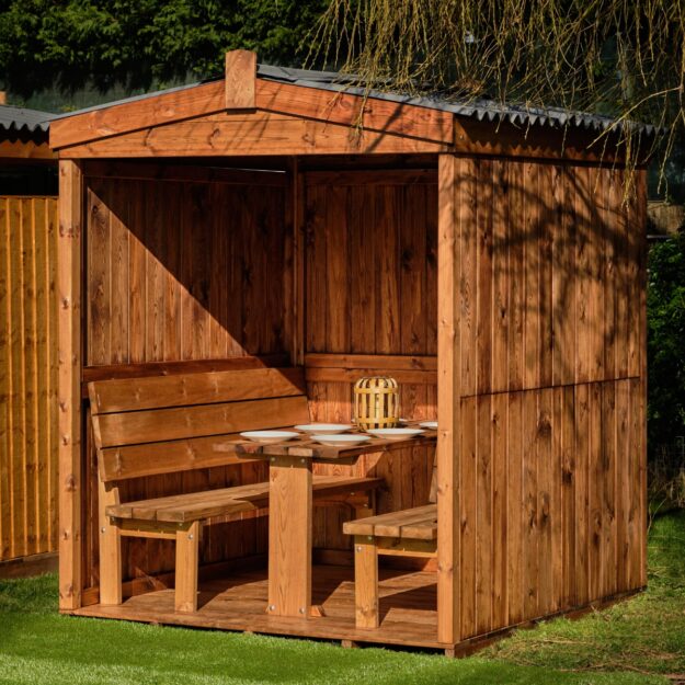 A 3 sided wooden dining cabin with a weather proof apex roof and fixed table and benches inside on grass