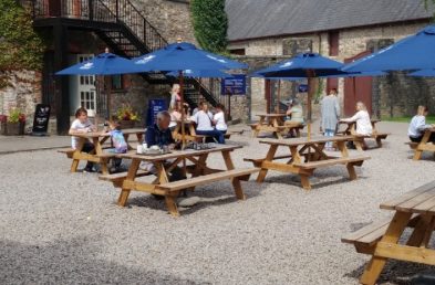 A gravel outdoor area with 8 wooden picnic tables spaced out with blue parasols and people sitting at them at a holiday park cafe