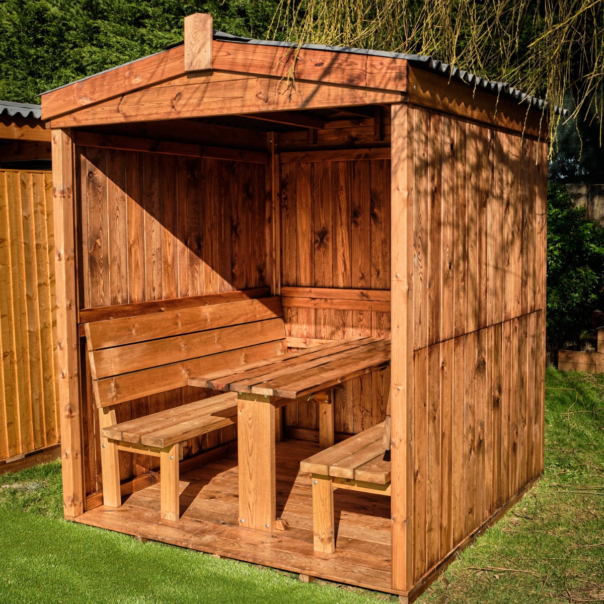 A wooden outdoor dining cabin, enclosed on 3 sides with an open side showing bench seats and a table seating up to 6 people the cabin has a pitched apex water proof roof