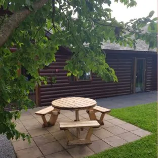 A circular 6 seater wooden picnic table on a patio of a wooden cabin at a holiday park