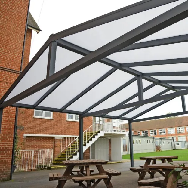 A grey metal framed gazebo with a polycarbonate apex roof on a school playground with picnic tables underneath it