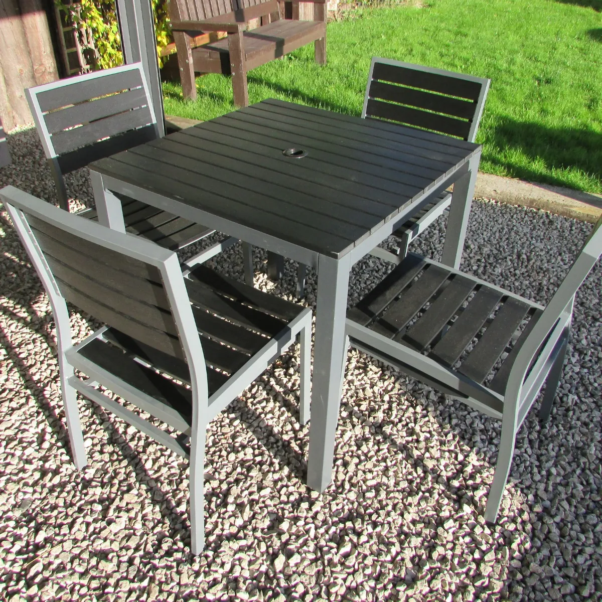 A square outdoor table with black plastic slats and a grey aluminium frame on a gravel patio with 4 matching diner chairs around it